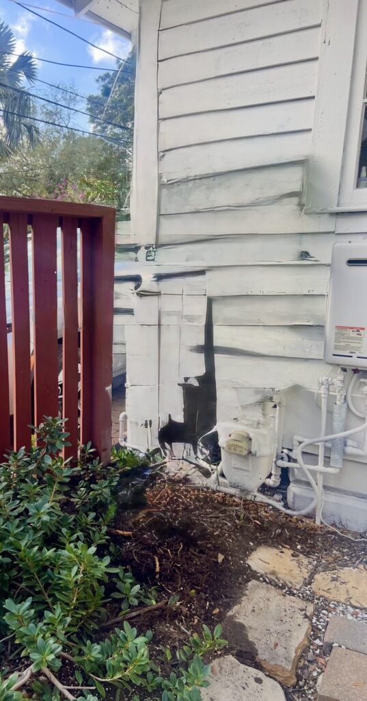 Damaged and peeling exterior wall with exposed pipes and a wooden fence nearby.